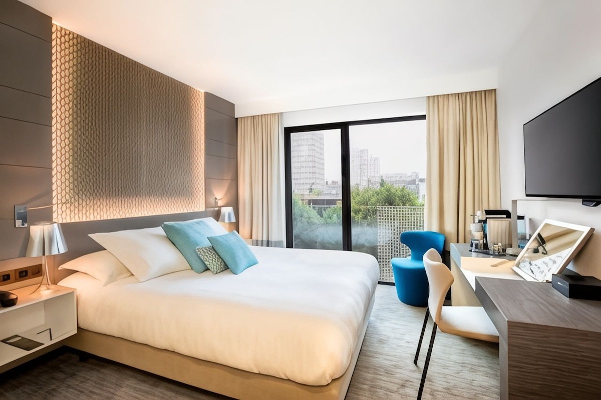 Habitación de hotel con una cama grande, un escritorio, una silla, un televisor y un balcón con vistas a la ciudad.