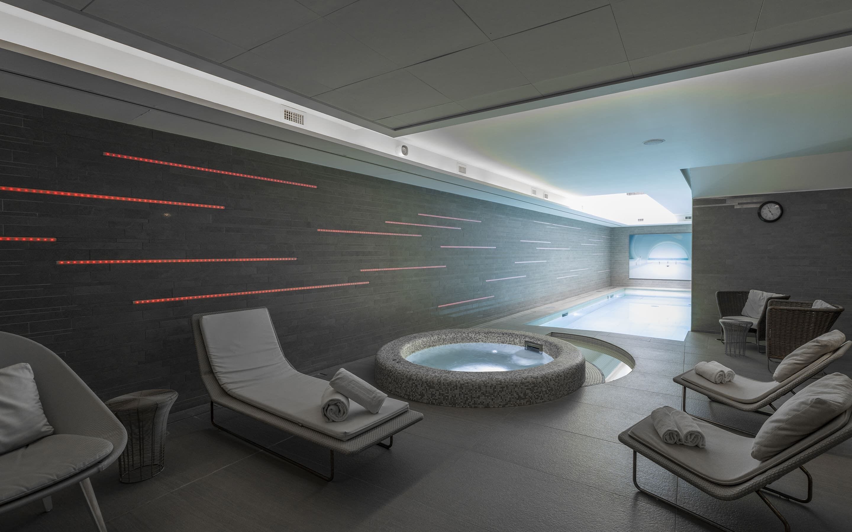 A modern indoor spa with a heated stone bath, lounge chairs, and a wall with red LED lighting.