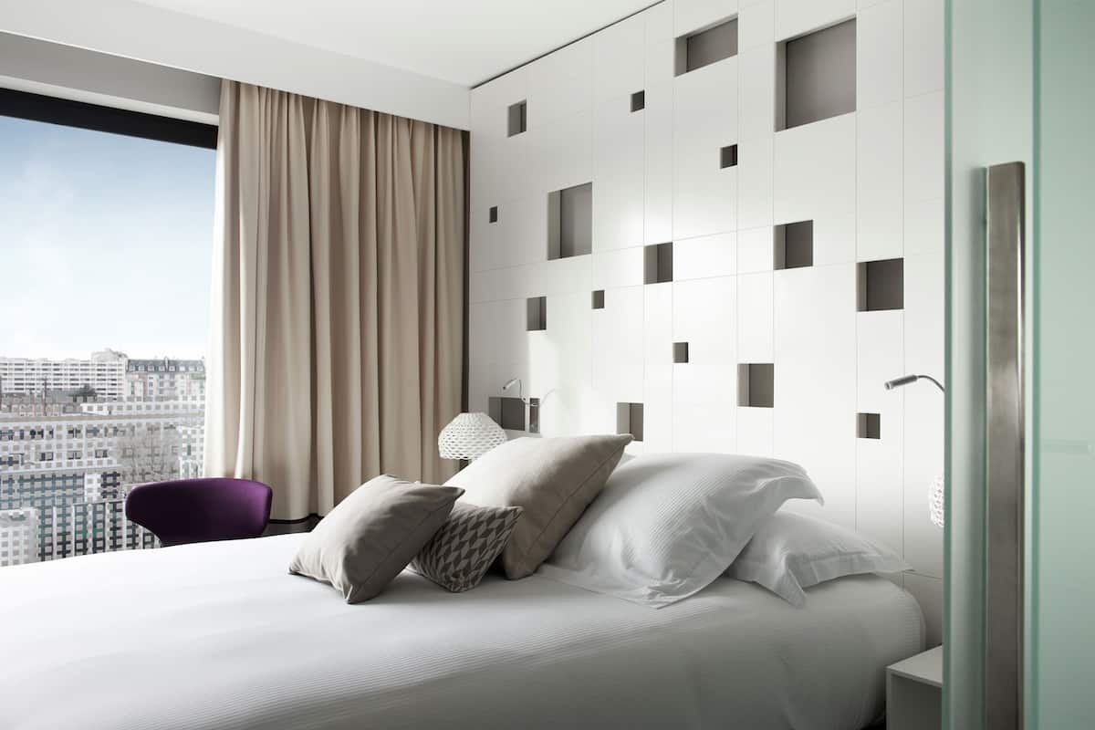 A modern hotel room with a large bed, a city view, and a minimalist wall design.