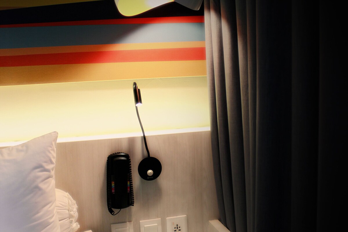 A hotel room with a bed, a bedside table, a lamp, a phone, and a wall with horizontal stripes.