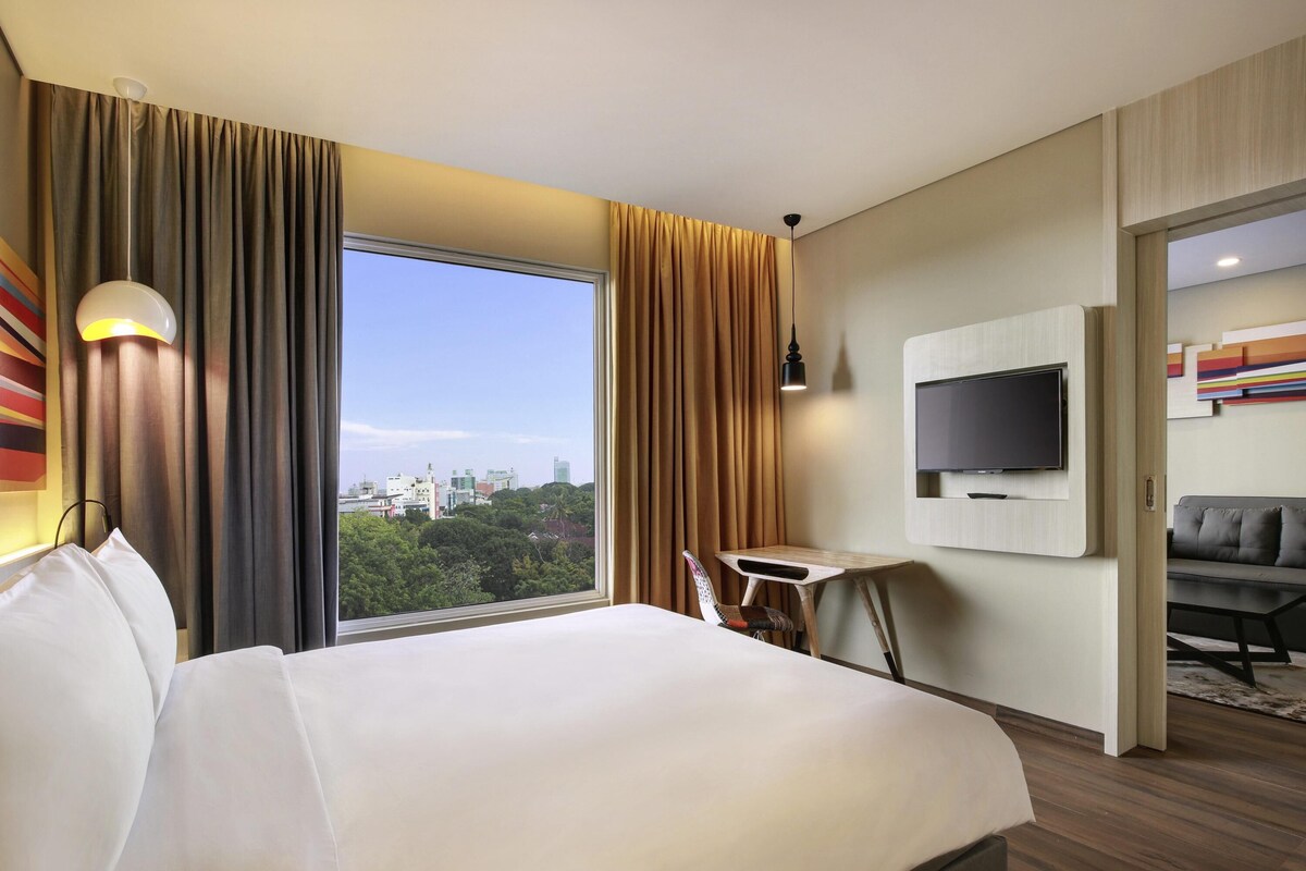 A hotel room with a large bed, a desk, a chair, a TV mounted on the wall, and a view of the cityscape through the window.