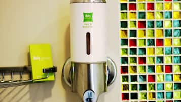 Dispensador de gel para cabello y cuerpo fijado a la pared, con etiqueta verde, junto a un muro de mosaico colorido.