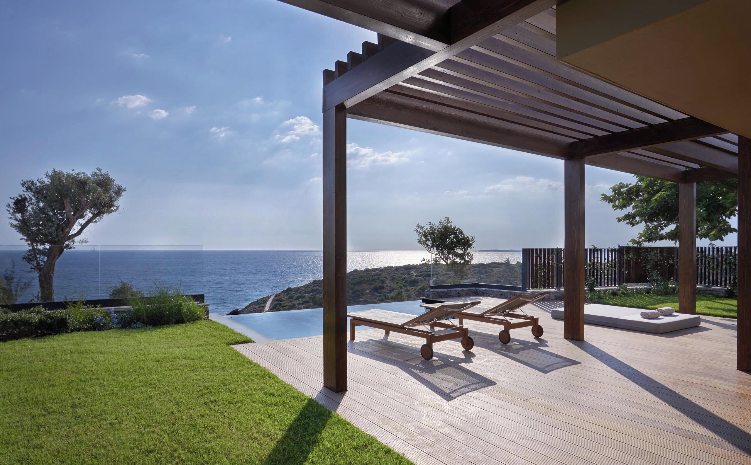 A wooden deck with a pergola, two lounge chairs, and an infinity pool overlooking the sea.