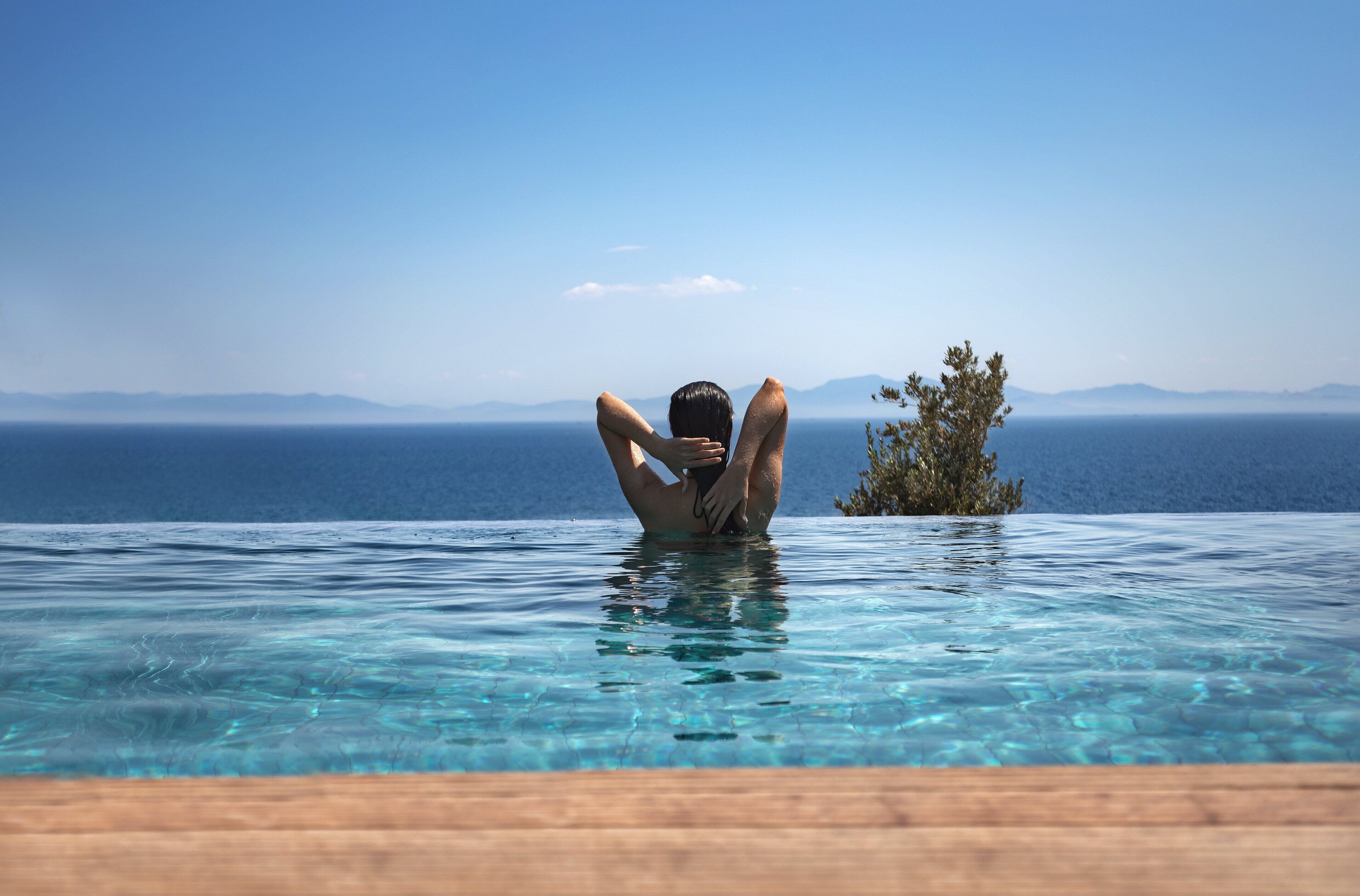 En person slapper af i en krystalklar pool med udsigt til havet og fjerne bjerge.