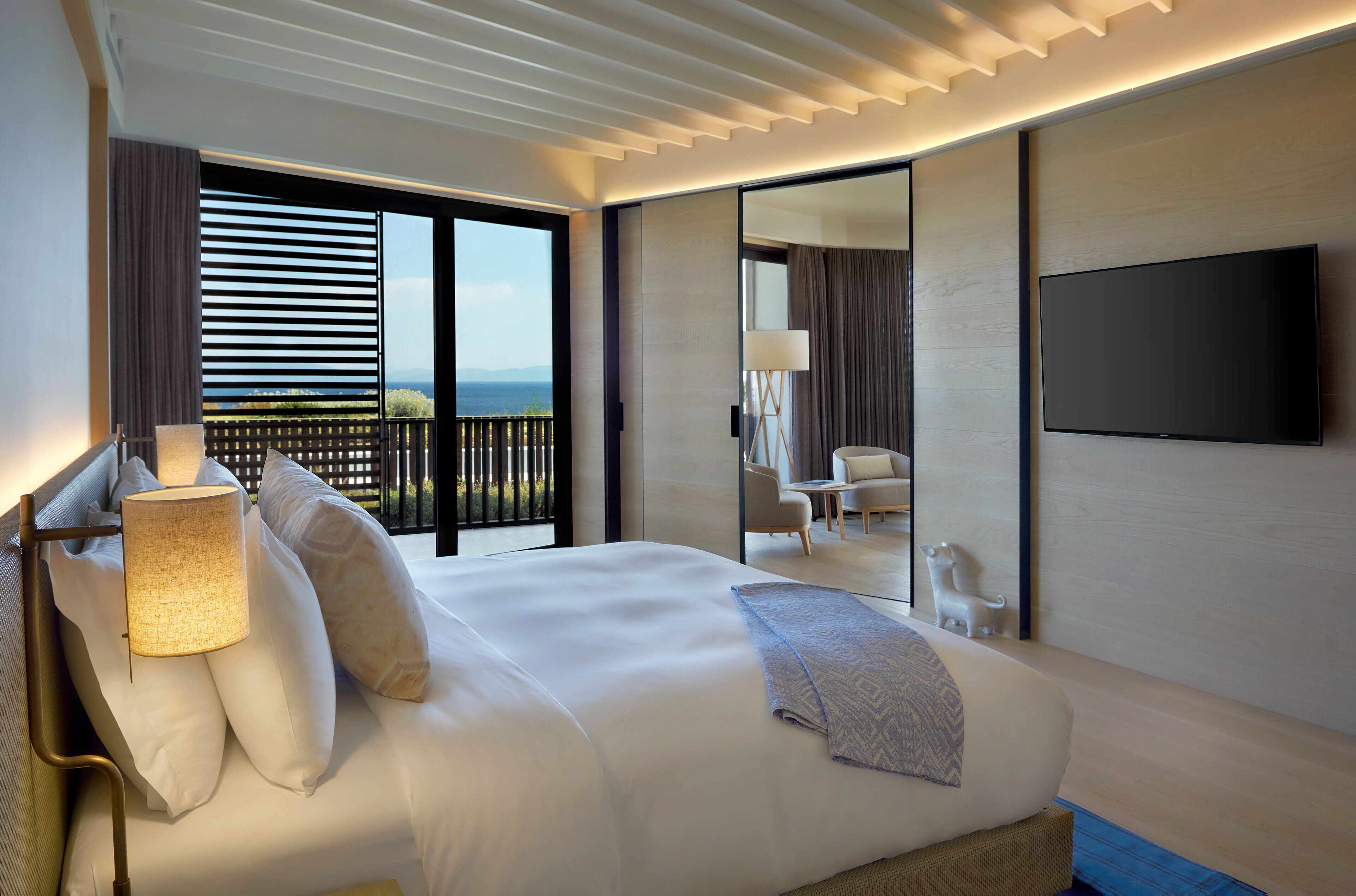 A modern hotel room with a large bed, a flat-screen TV, a balcony with a view, and a small seating area.