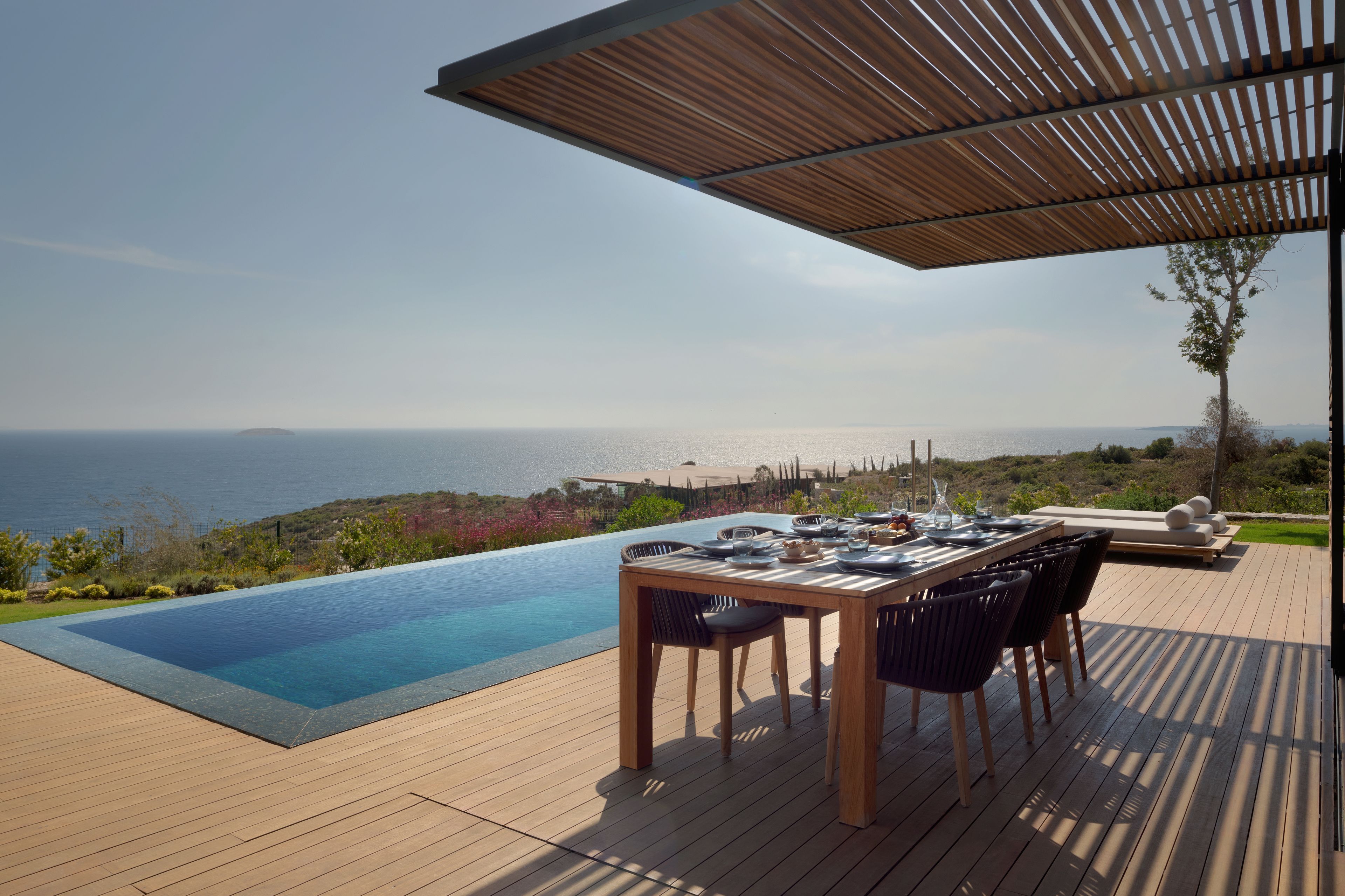 A wooden deck with a pool and a table set for dining, overlooking a scenic view of the sea.
