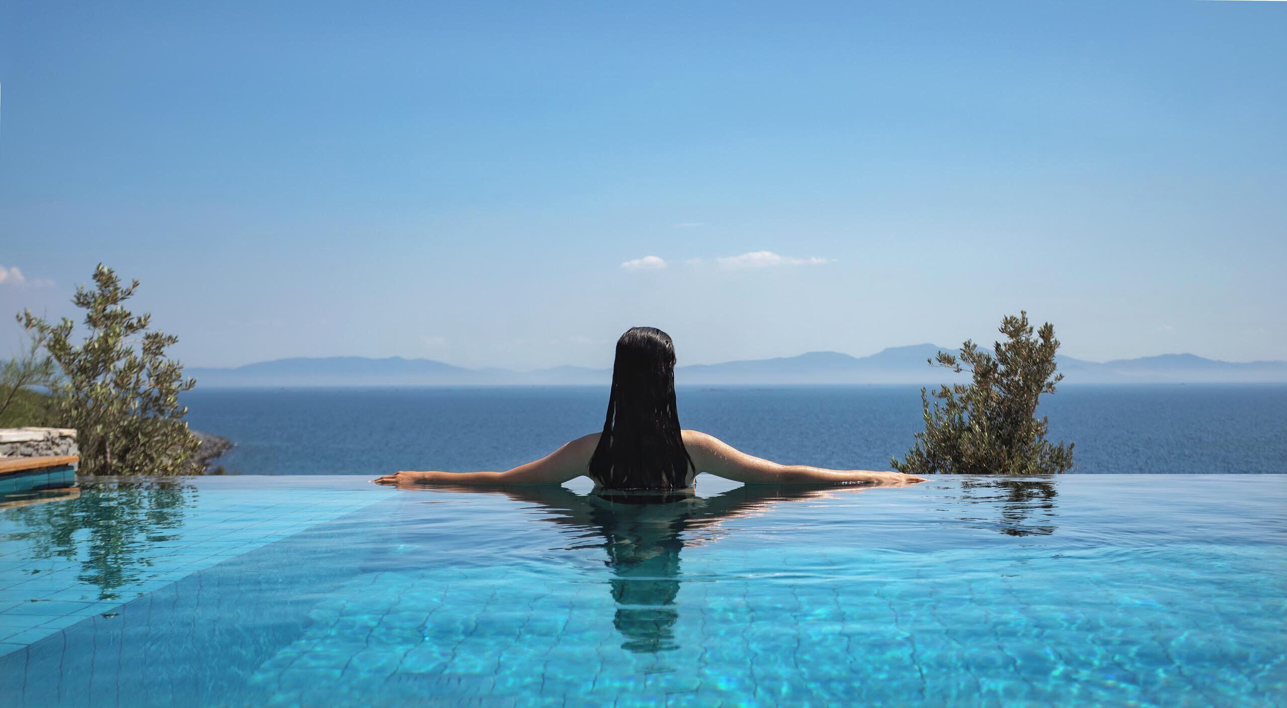 Una persona relajándose en una piscina infinita de color azul cristalino, con vistas al mar y montañas lejanas.