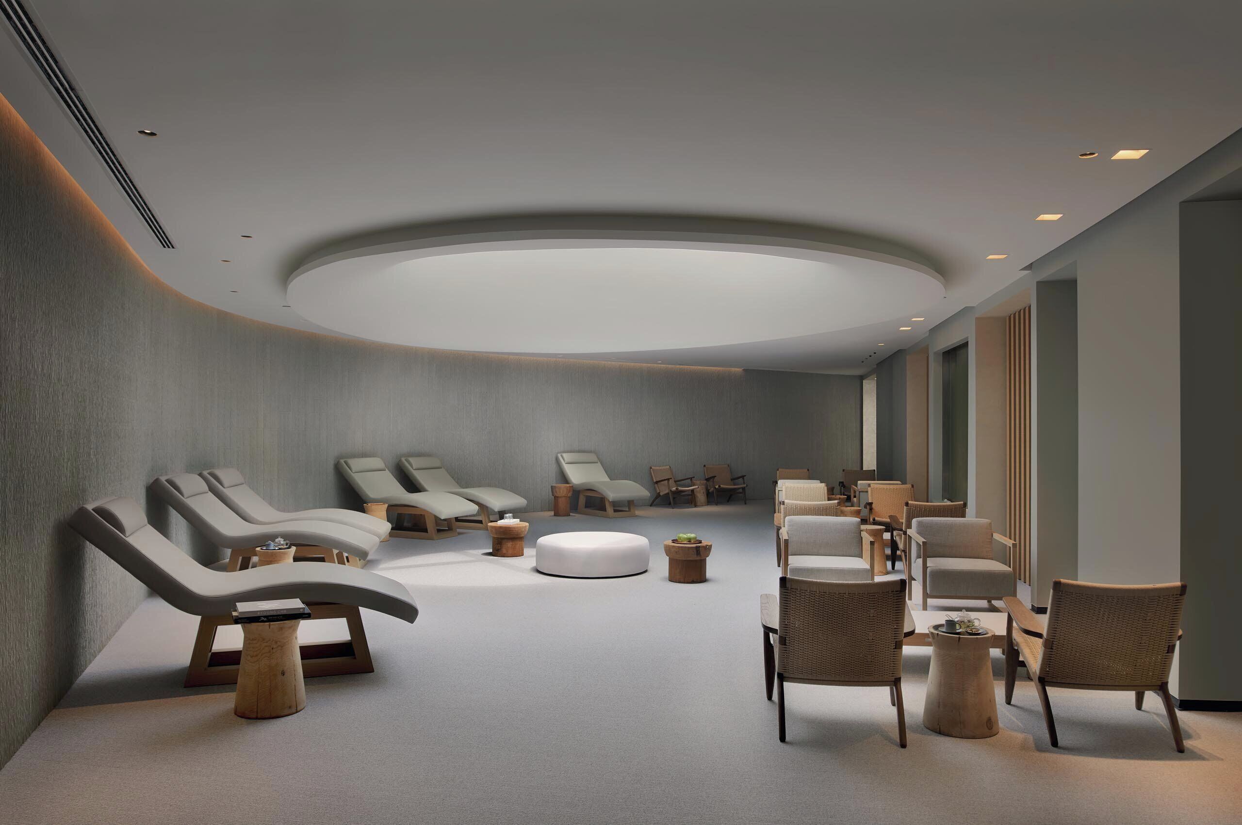 A modern spa room with multiple seating areas, including lounge chairs and wicker chairs, arranged around a central circular light fixture.
