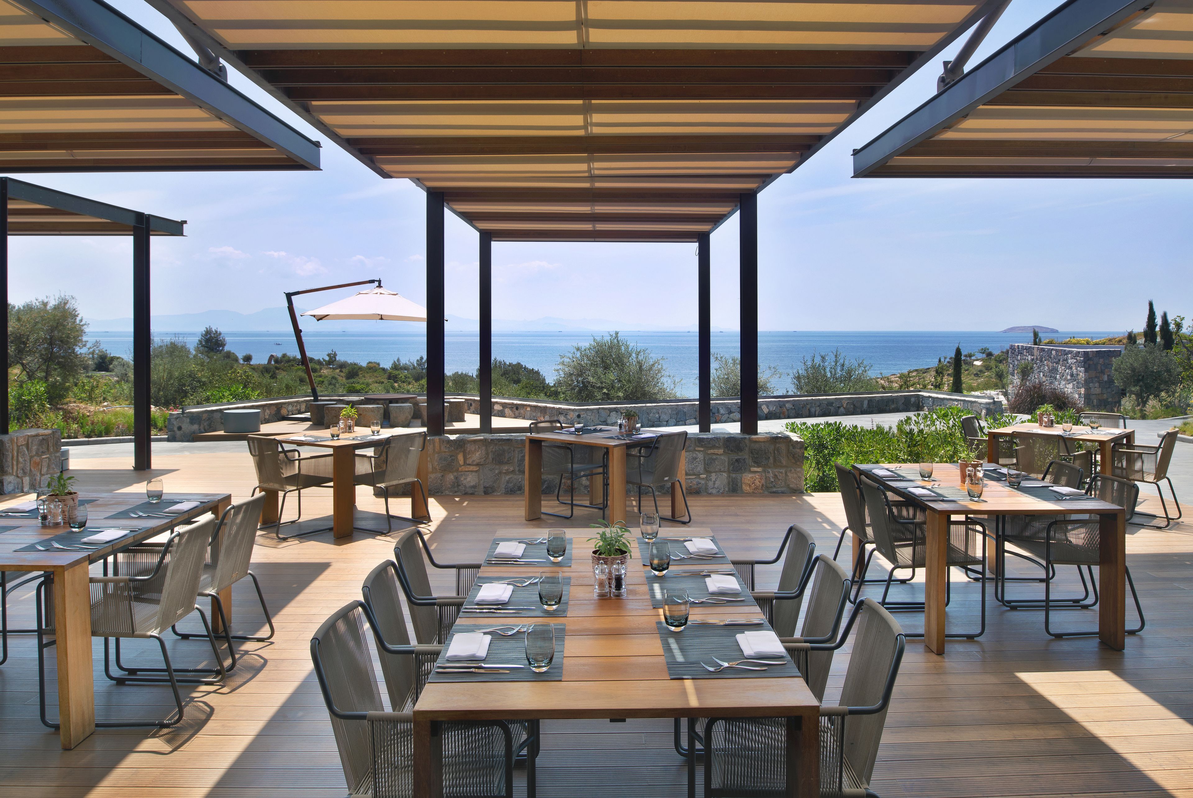Un área de comedor al aire libre con varias mesas y sillas, vista al mar y un muro de piedra al fondo.