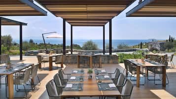 Un área de comedor al aire libre con varias mesas y sillas, vista al mar y un muro de piedra al fondo.