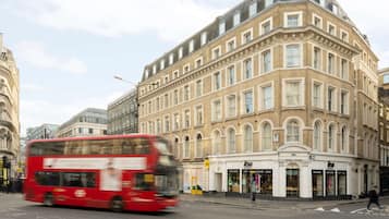A red double-decker bus on a city street, a corner building with multiple shops, and a clear sky.