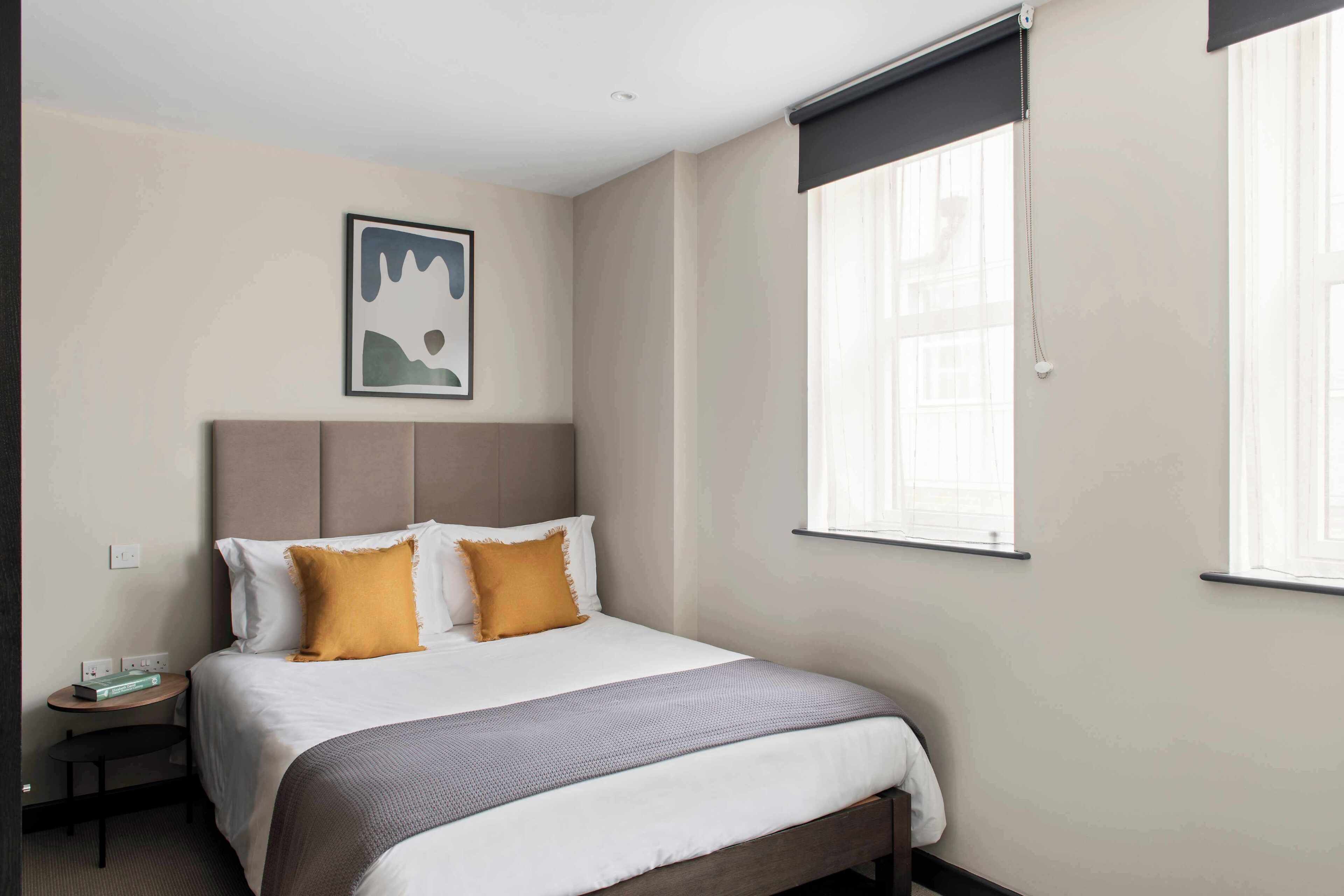 Ein Hotelzimmer mit Bett, Nachttisch, Wandbild und einem Fenster mit Jalousien.