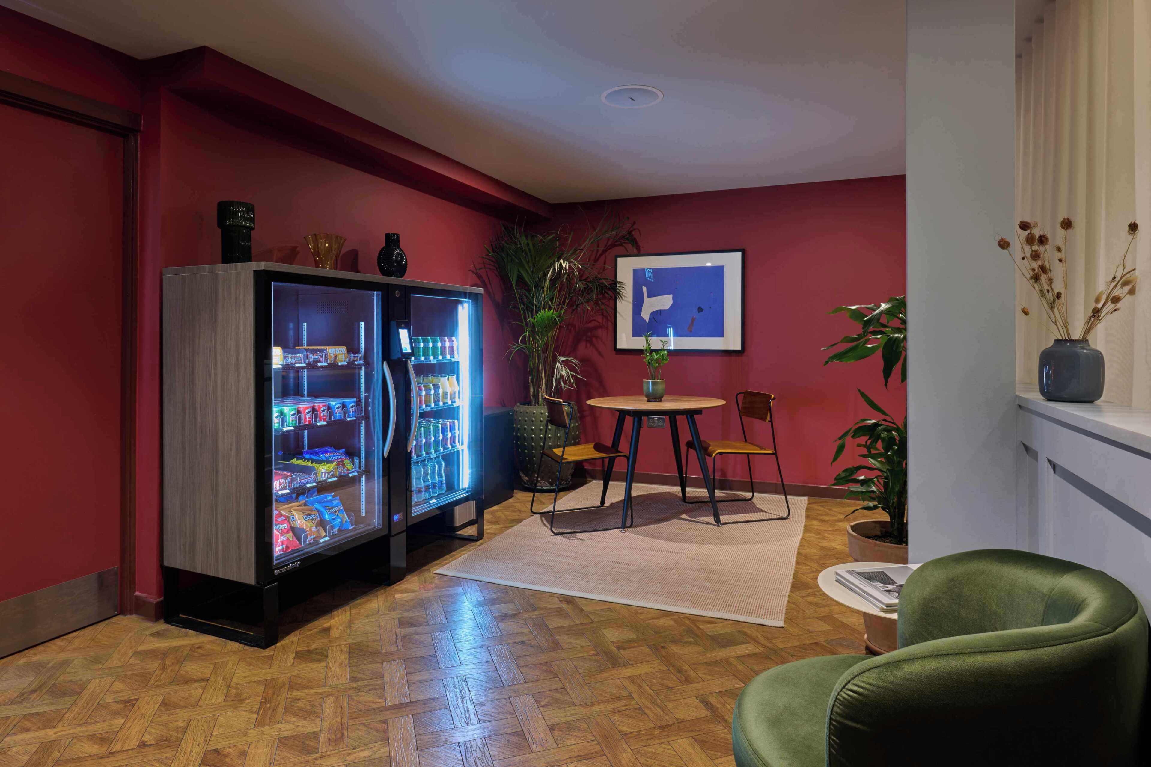 Une pièce avec un parquet en bois, un fauteuil vert, une petite table ronde, un réfrigérateur et une plante en pot.