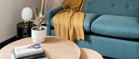A teal sofa with a mustard blanket, a small table with books, a potted plant, and a lamp.