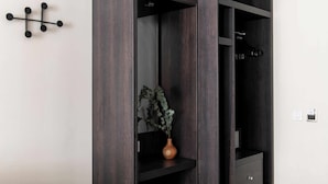 A dark wood wardrobe with a plant on the door, a black wall-mounted hook, and a small drawer. - Cove Cannon Street (London)