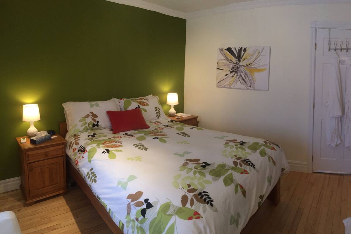 Un dormitorio con cama, mesitas de noche, lámpara, una puerta con toallas y un cuadro en la pared.