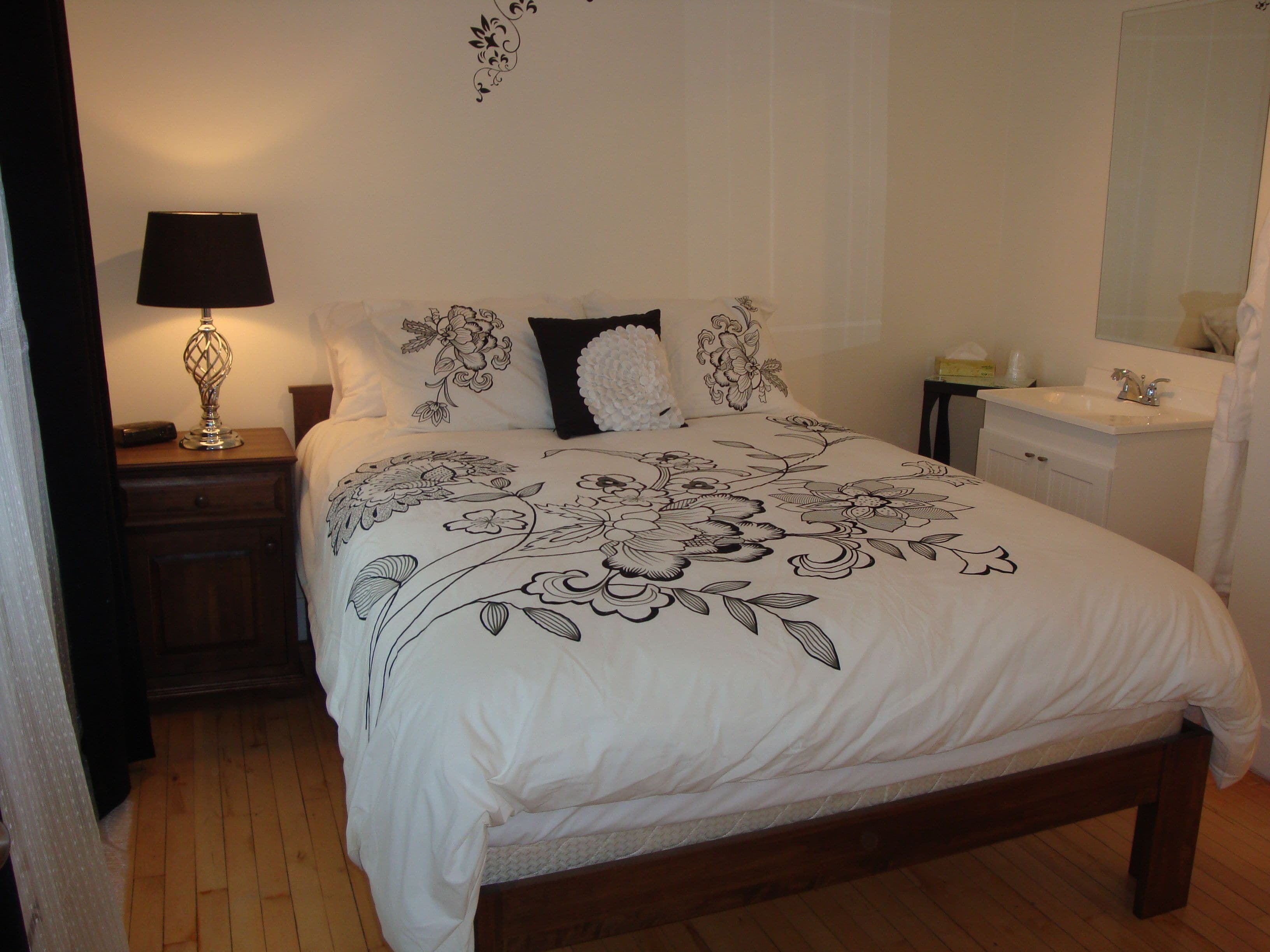 A neatly made bed with a floral bedspread, a bedside lamp, a nightstand, a dresser, and a bathroom with a mirror and sink.