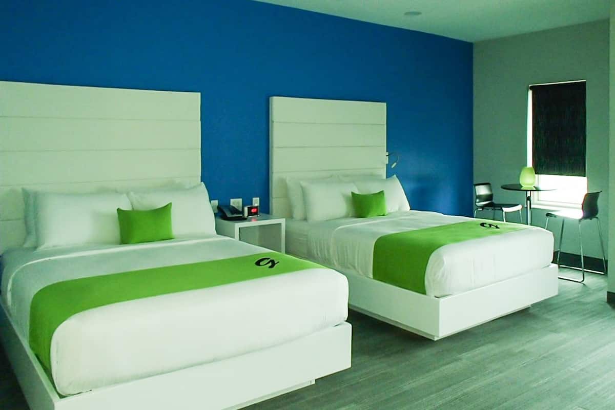 A hotel room with two beds, each with white bedding and green accents, a blue accent wall, and a small desk with a chair.