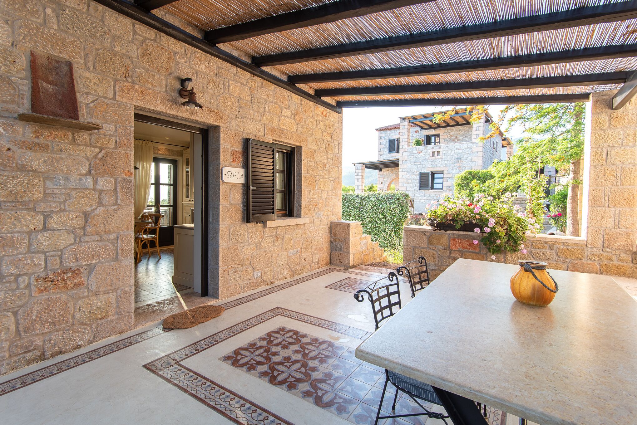 A stone-walled room with a tiled floor, a table with chairs, a window with shutters, and a view of buildings in the background.