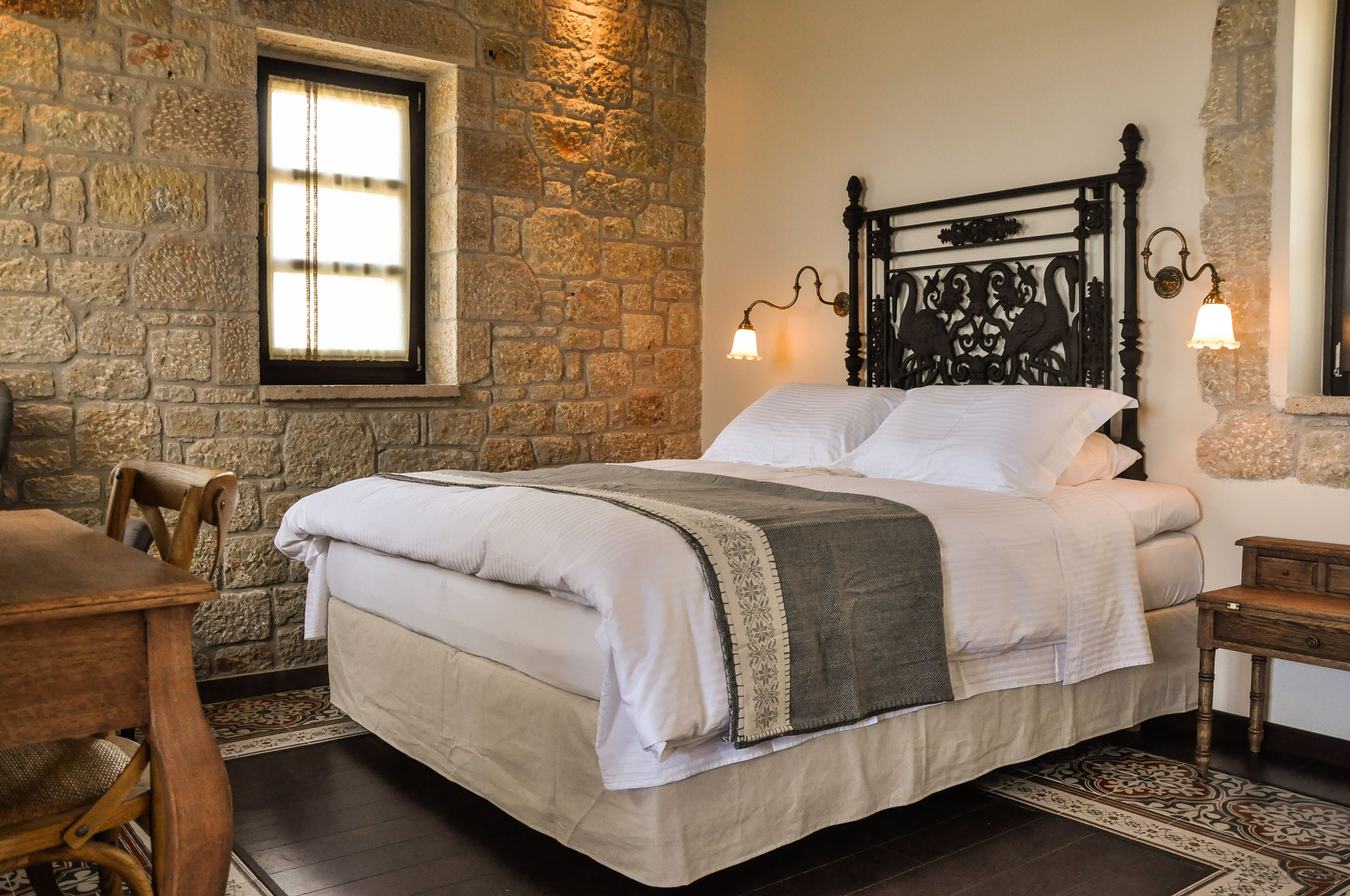 A neatly made bed with a headboard, a wooden nightstand, and a stone wall with a window.