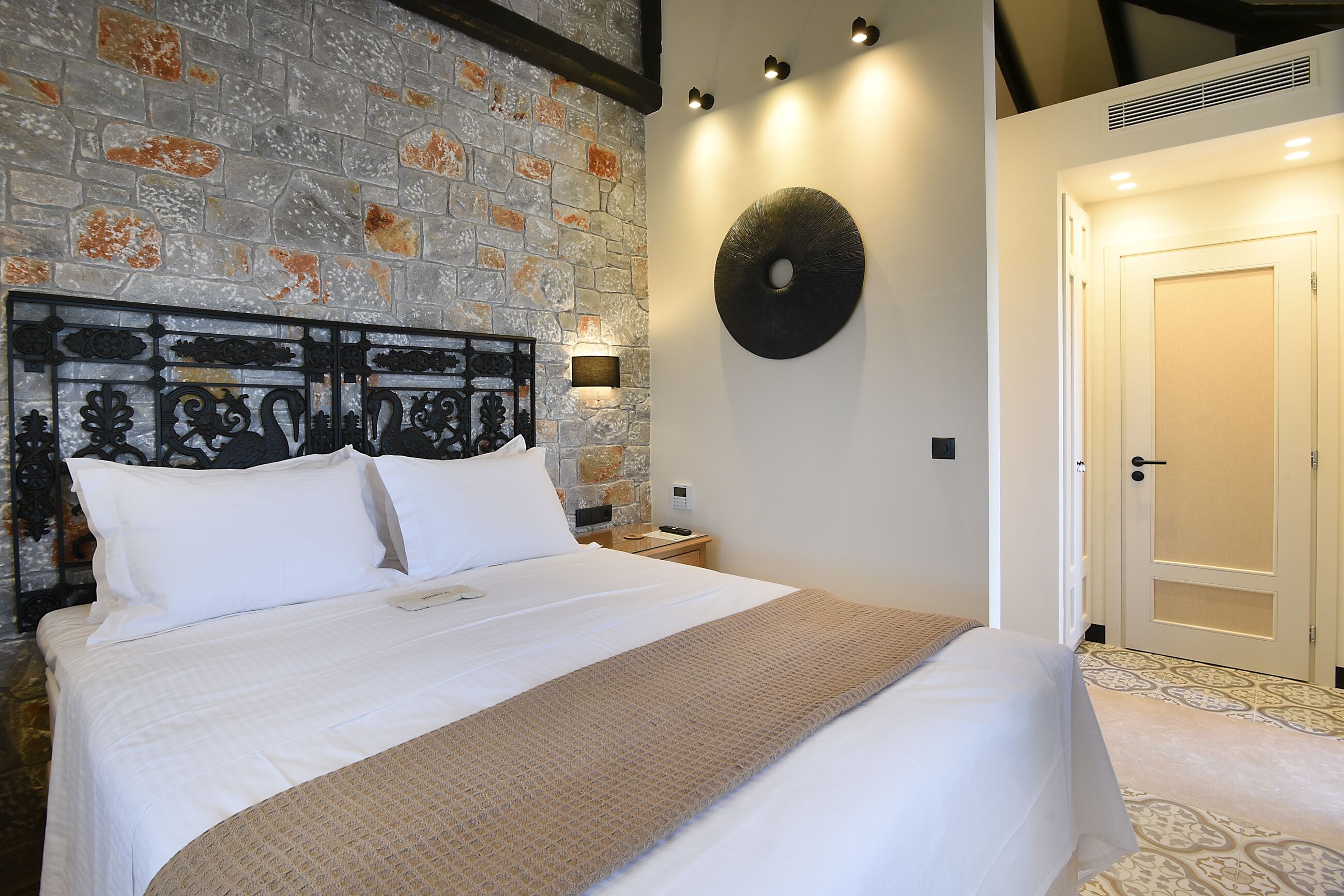 Una cama bien tendida con sábanas blancas y una manta marrón, una pared de piedra con un cabecero metálico decorativo, una obra de arte circular colgada en la pared y una puerta cerrada con interruptor de luz.