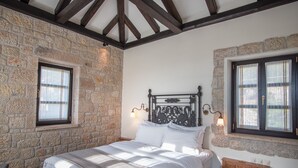 A bedroom with a stone wall, a wooden ceiling, a bed with white bedding, and two windows with curtains. - Polismata - Private Residences (West Mani)