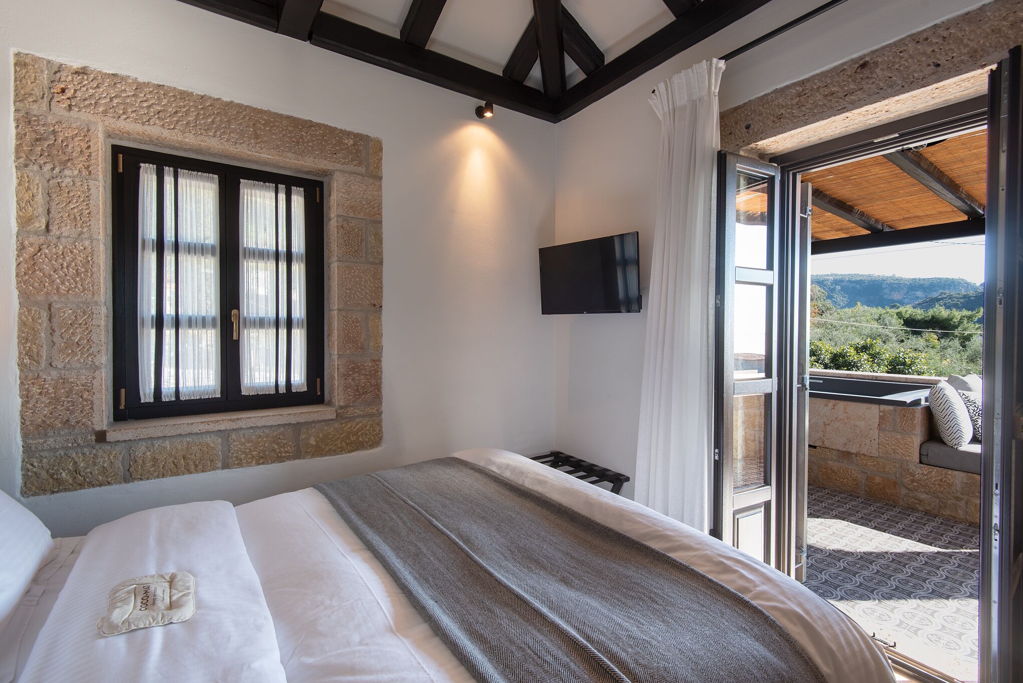 Een slaapkamer met een stenen muur, een bed met dekbed, een aan de muur bevestigde televisie en een balkon met uitzicht.