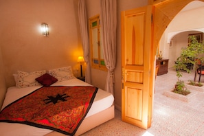 A bedroom with a bed, a nightstand, a lamp, a window with curtains, and an open door leading to a courtyard with plants. - Riad Inaka (Marrakech)