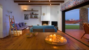 A modern living room with a fireplace, wooden beams, a sofa, a coffee table with candles, and a view of the outside.