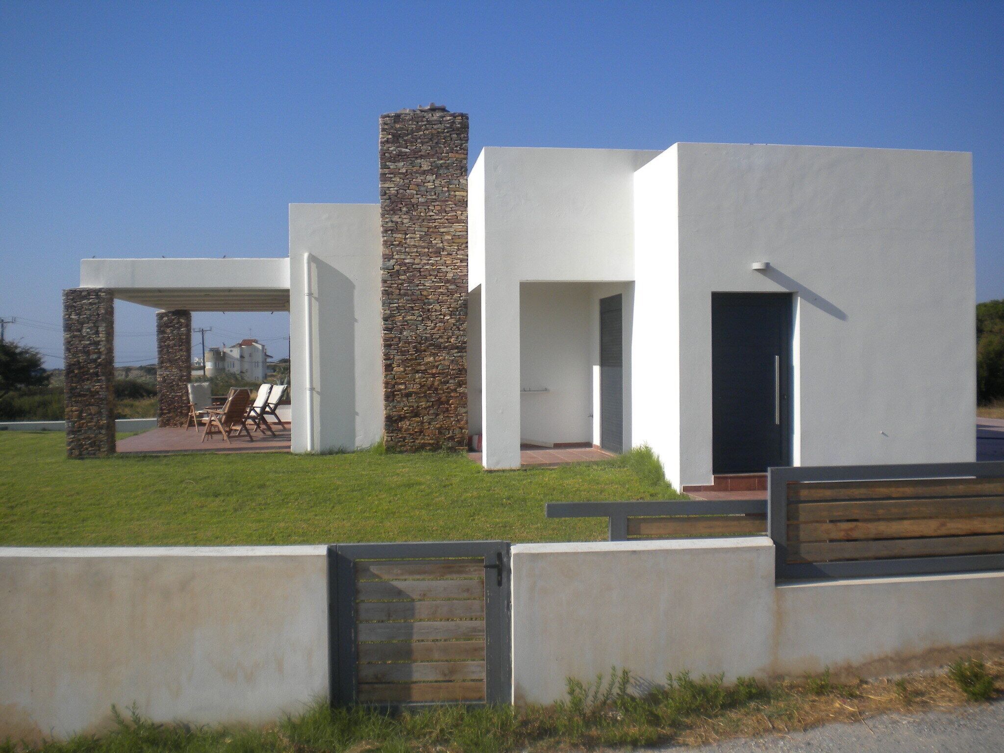 Una casa moderna con chimenea de piedra, un porche cubierto con sillas y una entrada protegida por un portón.