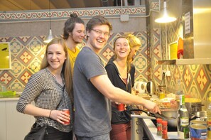 A group of people in a kitchen area with a patterned backsplash, a counter with food items, and a sign instructing to use the kitchen hood. - Bed & Bike Hostel Barcelona (Barcelona)