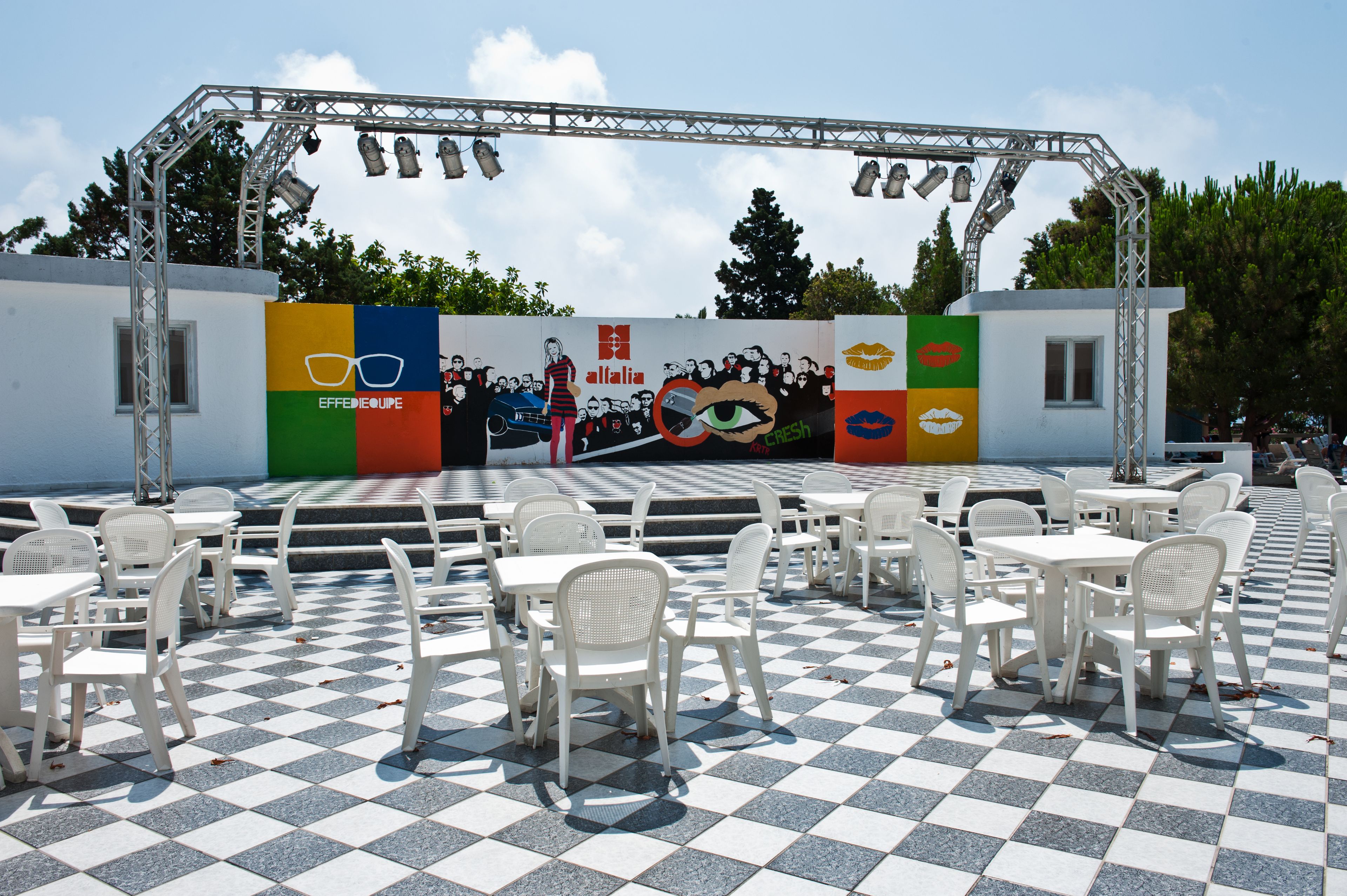 An outdoor seating area with white plastic chairs and tables, a stage with a colorful mural, and a metal truss overhead.