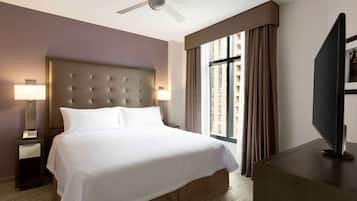 Habitación de hotel con una cama grande, ventilador de techo, televisor sobre un soporte de madera y una ventana con cortinas.