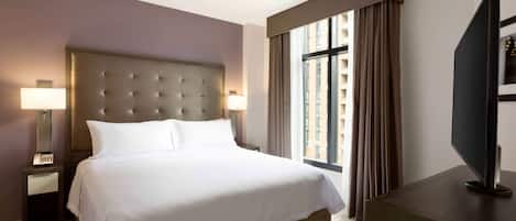Habitación de hotel con una cama grande, ventilador de techo, televisor sobre un soporte de madera y una ventana con cortinas.