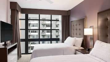 A hotel room with two beds, a TV, a ceiling fan, a large window with a view of buildings, and a nightstand with a lamp.