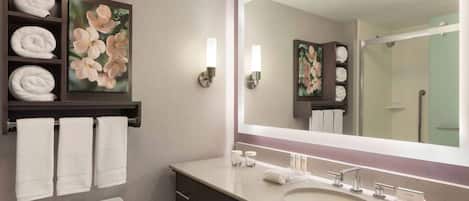 A bathroom with a toilet, sink, and mirror. There are towels on a shelf and a shower visible in the mirror.