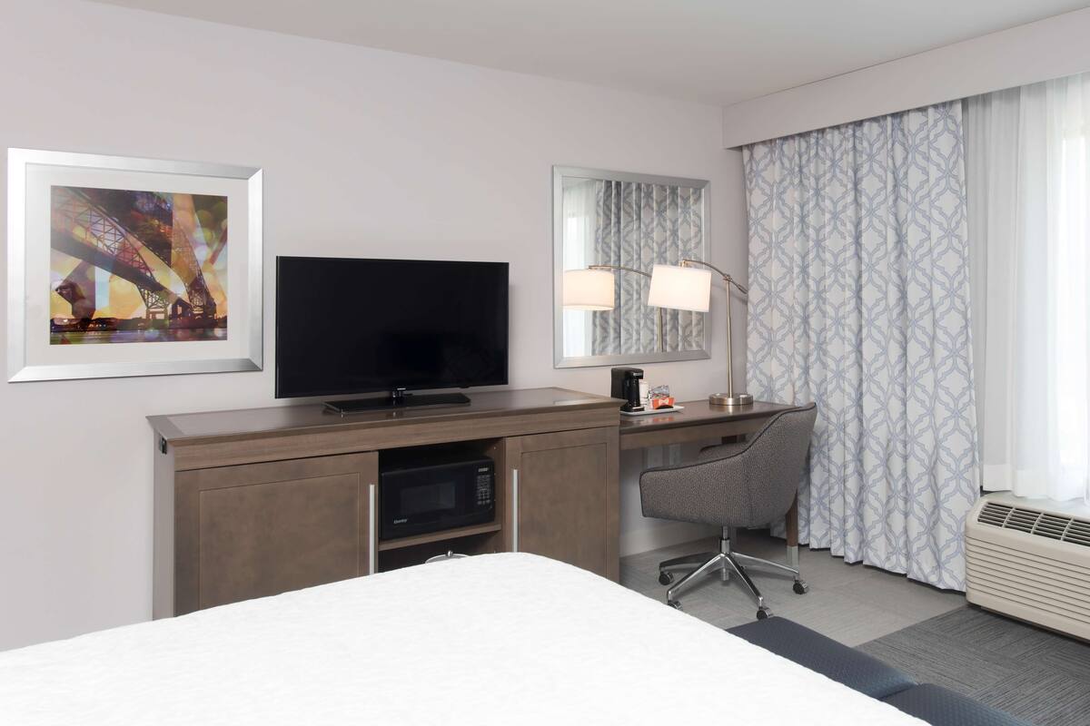 A hotel room with a bed, a desk with a computer, a flat-screen TV, a microwave, a chair, and a large window with curtains.