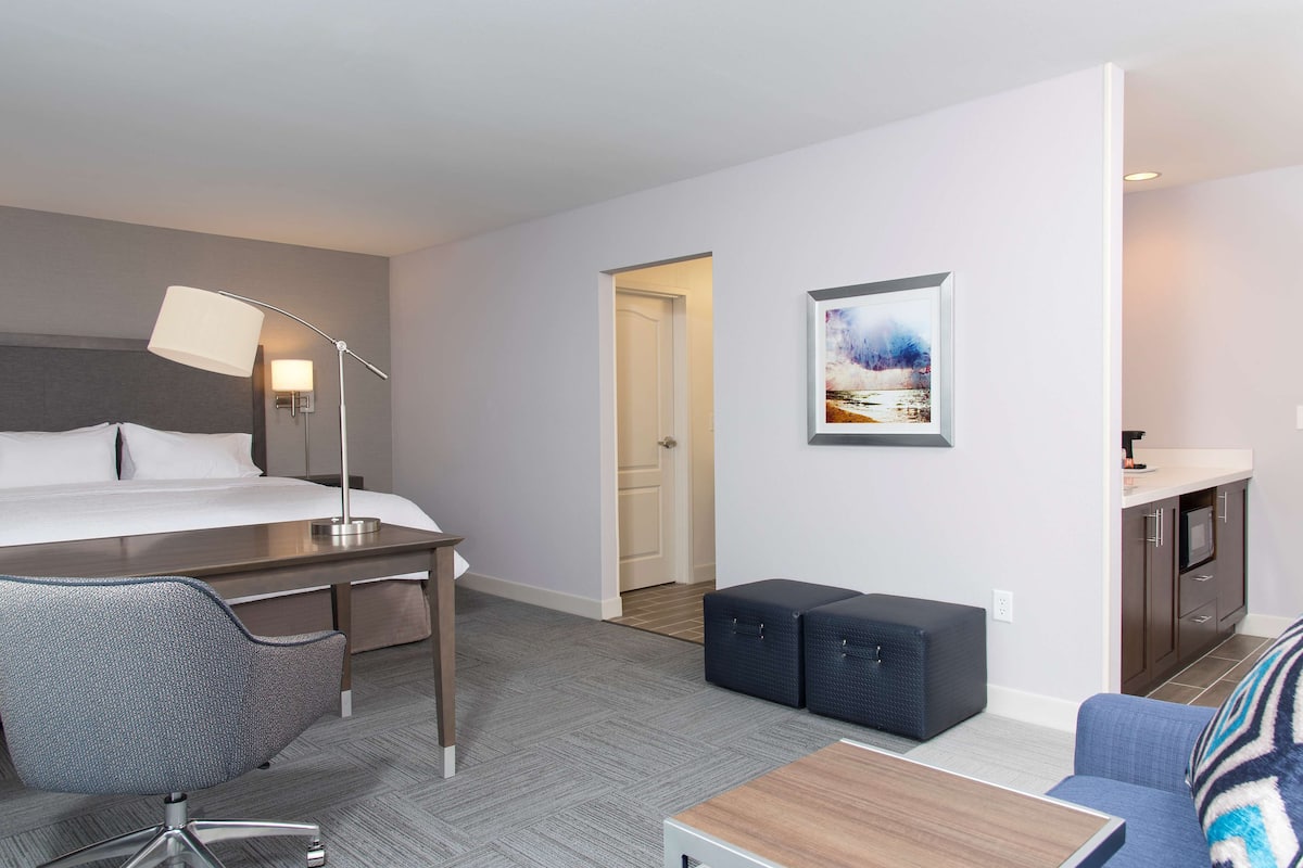 A hotel room with a bed, desk, chair, kitchenette, and a painting on the wall.