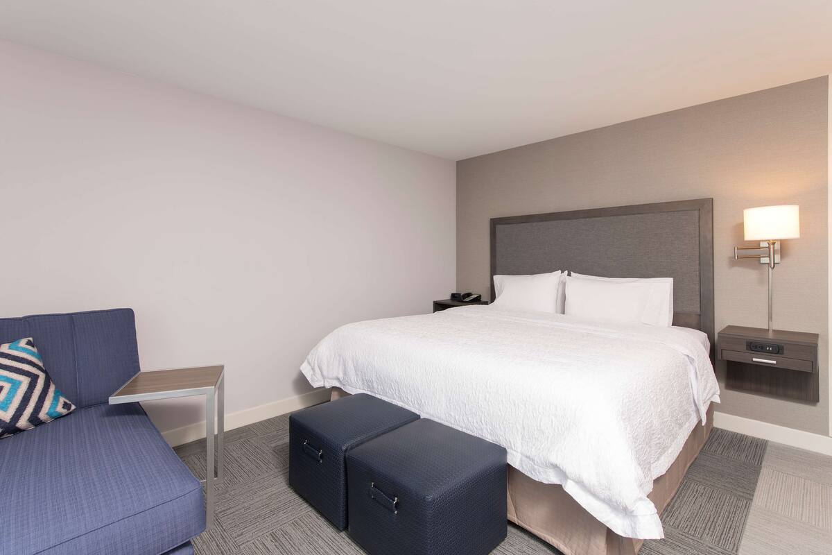 A hotel room with a bed, a small bedside table, a nightstand, a lamp, a sofa, and two ottomans.