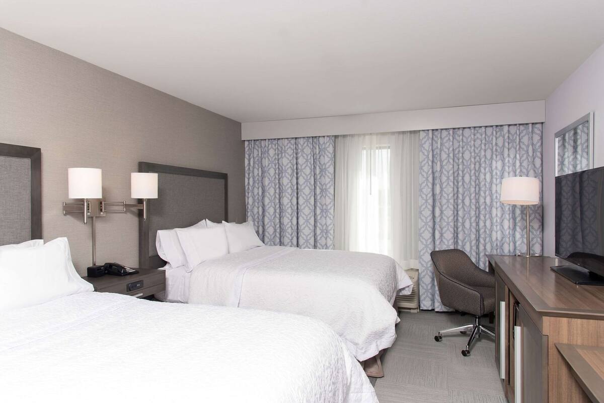 A hotel room with two beds, a desk, a chair, a TV, and a lamp.