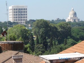 A view of a cityscape with a large white building with multiple windows, a dome-shaped structure, and dense green trees in the foreground. - Attico dei Pini (Rome)