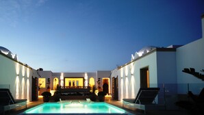 Un espace piscine avec des chaises longues et une vue sur un bâtiment doté d’arcades et d’un éclairage décoratif.