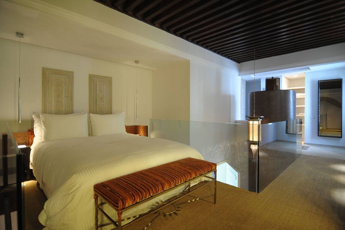 A bedroom with a large bed, a bench, a bedside table, and a mirror.