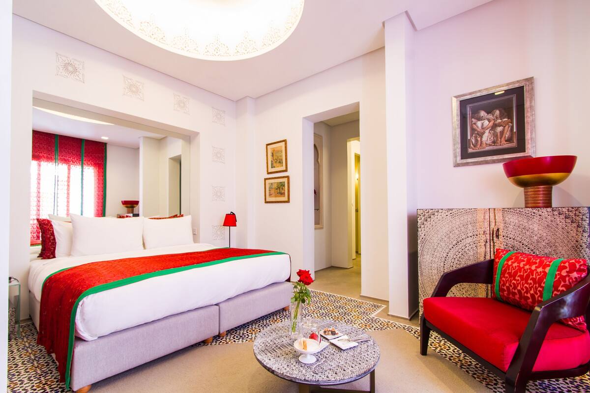 A hotel room with a bed, a sofa, a round table with a vase and a red cushion, and a framed picture on the wall.