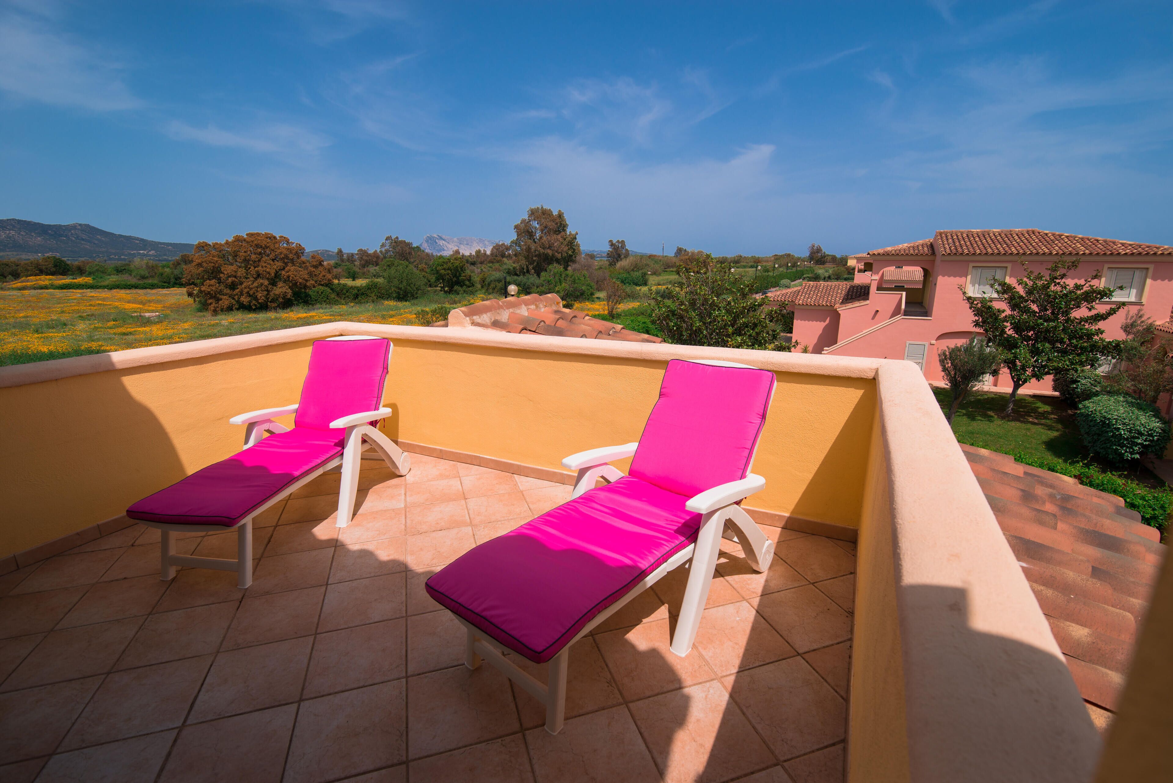 Une terrasse avec deux fauteuils blancs, dotés de coussins roses, un mur jaune et une vue sur un bâtiment rose et un paysage verdoyant.