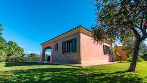 Un pequeño edificio rosa con fachada de piedra y contraventanas verdes, rodeado de un césped cuidado y árboles.