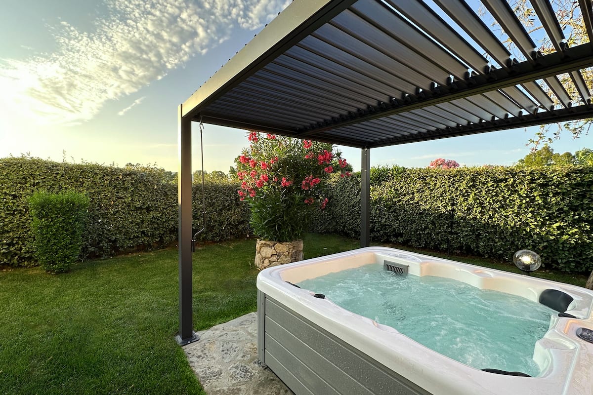 A hot tub under a pergola in a garden with a hedge and flowering plants.