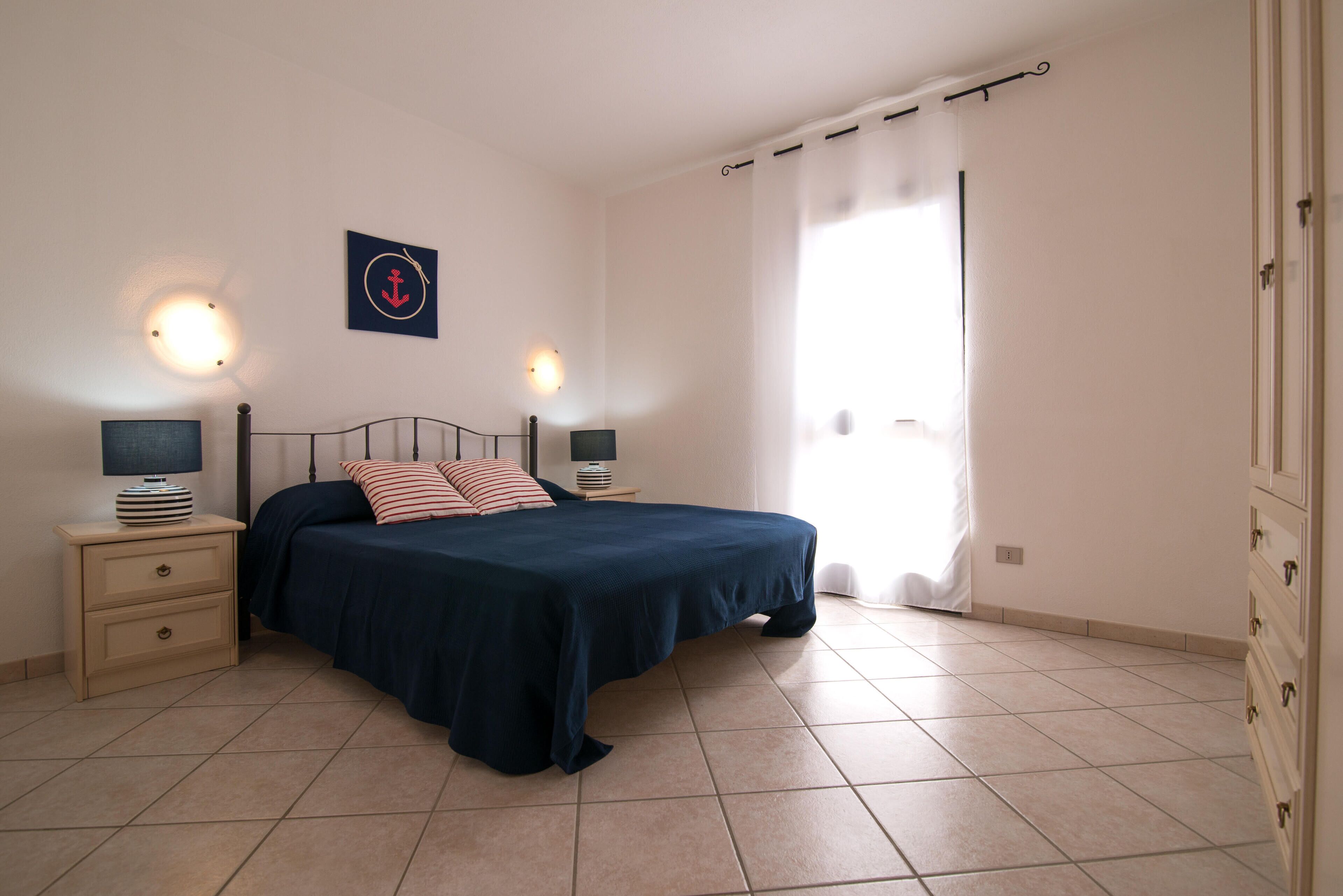 Un dormitori amb llit, tauletes de nit, una làmpada penjada a la paret i una finestra amb cortines.