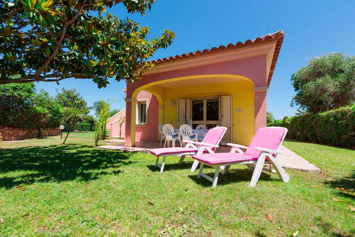 A small house with a patio, two pink lounge chairs, and a table with chairs.