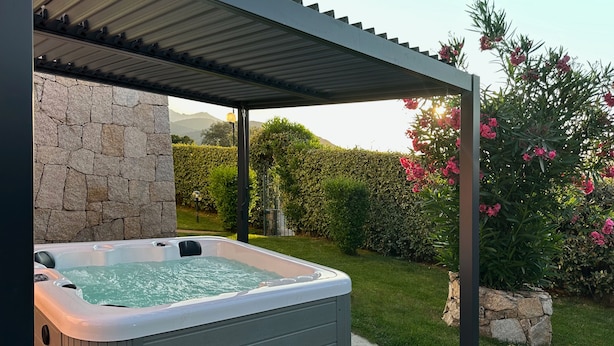 Un jacuzzi bajo una pérgola con paredes de piedra y un jardín con flores y setos.